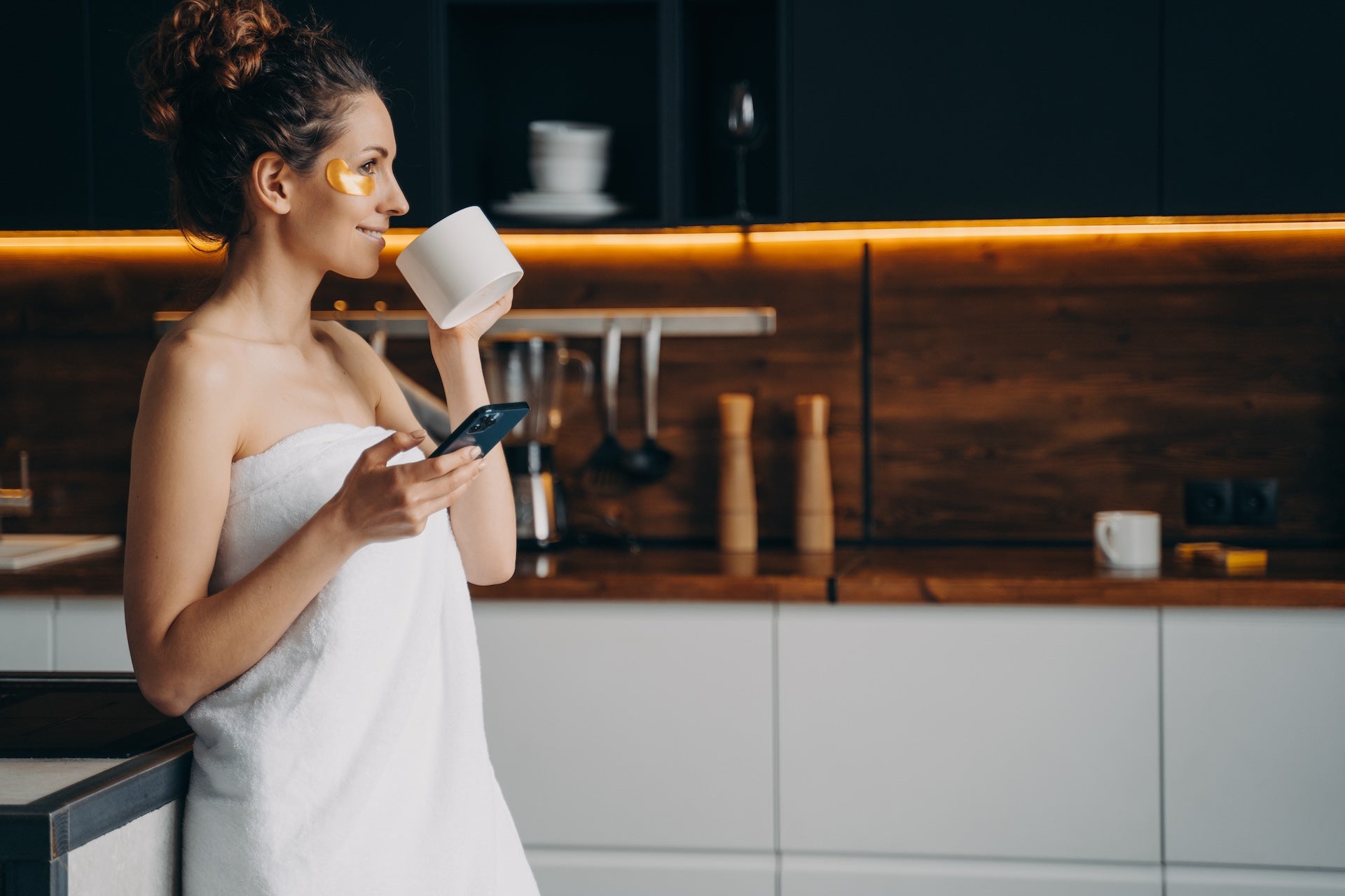 Lady wrapped in a towel with a beauty patch beside her eye checking her phone drinking coffee with collagen powder in her kitchen.