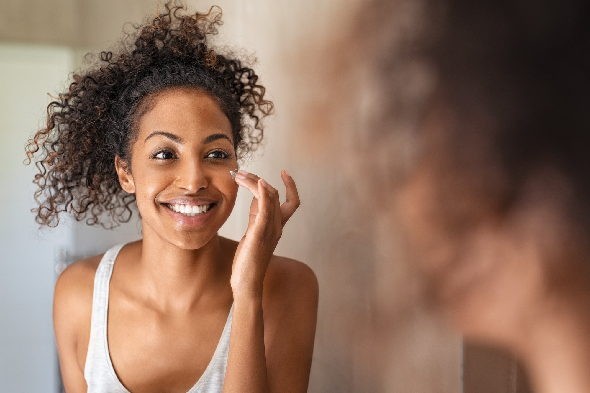 Lady with curly black hair looking in the mirror at her rejuvenated skin tone after using liquid collagen drink